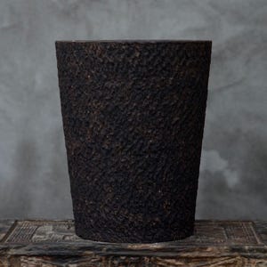Puede incluir: Un jarrón cilíndrico marrón oscuro y texturizado con una forma ligeramente cónica. La superficie tiene un aspecto rugoso, similar a la corteza. El jarrón se exhibe sobre una superficie de madera sobre un fondo gris. Este jarrón decorativo es adecuado para la decoración del hogar.
