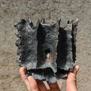 Op de afbeelding: Een donkergrijze, getextureerde, abstracte sculptuur met een ruw, ongelijkmatig oppervlak. De sculptuur heeft een rechthoekige vorm met verticale ribbels en een kleine, rechthoekige opening. Het object wordt in een hand gehouden.