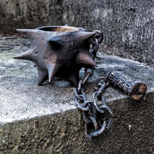 Puede incluir: Una maza metálica oscura con protuberancias puntiagudas, conectada por una cadena a un mango cilíndrico. El arma descansa sobre una superficie de piedra gris y desgastada, evocando un tema medieval o de fantasía.