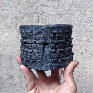 Puede incluir: Una maceta cilíndrica gris oscuro con un diseño texturizado y en capas. La maceta tiene una apariencia rugosa, similar a la piedra, con bandas horizontales de diferentes anchos. La estética general es rústica y moderna.