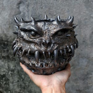 Pode incluir: Uma tigela escura e metálica com um rosto monstruoso esculpido no exterior. Possui dentes afiados, chifres e espinhos ao redor da borda, criando uma aparência ameaçadora. A cor é um bronze escuro e envelhecido. A tigela é segurada em uma mão.