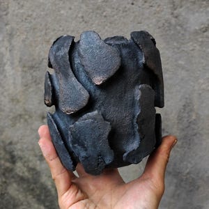 Könnte beinhalten: Eine dunkelgraue Keramikvase mit einer strukturierten, organischen Form. Die Vase hat blütenblattartige Vorsprünge an der Außenseite. Die handgefertigte Keramik hat eine raue, matte Oberfläche. Die Vase wird von einer Hand vor grauem Hintergrund gehalten.