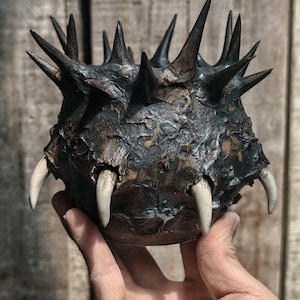 May include: A dark, textured sculpture resembling a spiky orb, with sharp, pointed protrusions and several large, ivory-colored teeth. The sculpture has a rough, weathered appearance, with a dark brown and black color palette.