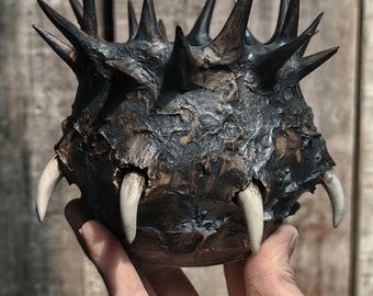 May include: A dark, textured sculpture resembling a spiky orb, with sharp, pointed protrusions and several large, ivory-colored teeth. The sculpture has a rough, weathered appearance, with a dark brown and black color palette.