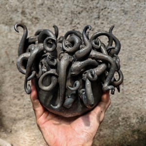 May include: A dark gray metal sculpture of a bowl with numerous curled tentacles extending outward. The tentacles have a textured surface and are arranged around the bowl's rim. The sculpture is held in a hand.