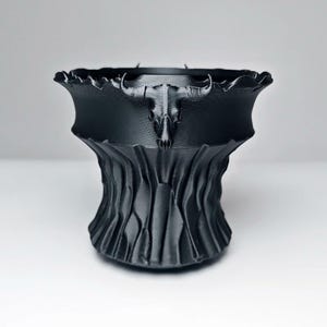 Peut inclure: Un pot décoratif noir au design unique. Le pot présente une base texturée en forme d'arbre et un dessus avec un crâne de taureau stylisé. Le pot est fait d'un matériau sombre et solide et convient à une utilisation intérieure ou extérieure.