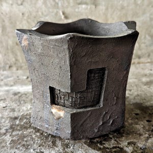 Puede incluir: Un jarrón de cerámica artesanal gris oscuro con un diseño angular único. El jarrón presenta una superficie texturizada y un recorte rectangular que revela un patrón interior similar a un ladrillo. El borde superior tiene un contorno ondulado.