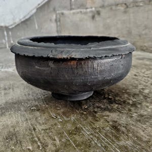 May include: A dark gray, handcrafted ceramic bowl with a wide, textured rim and a rounded body. The bowl sits on a small, circular base. The surface has visible brushstrokes, giving it a rustic appearance. The bowl is suitable for decorative or functional use.