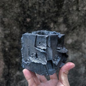 Può includere: Un vaso quadrato grigio scuro e strutturato con una superficie ruvida e irregolare. Il vaso è tenuto in mano, mostrando il suo aspetto unico, quasi roccioso. Progettato per piccole piante o succulente.