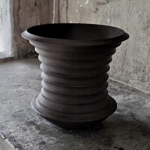 May include: A dark brown, matte-finished ceramic vessel with a wide, flared rim and a series of horizontal, raised ridges encircling the body. The object features a tapered, hourglass-like silhouette with a rounded base and a wide, open top.