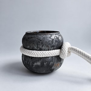 May include: A dark, mottled charcoal-coloured ceramic vessel with a rounded, bulbous shape. A thick, white braided rope is wrapped tightly around the centre of the vessel, secured with a prominent knot on the right side of the object.
