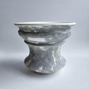 May include: A ceramic bowl with a wide, flared rim and a tapered, hourglass-shaped body. The surface features a mottled, textured finish in shades of slate grey and white, creating a marbled appearance across the entire exterior.