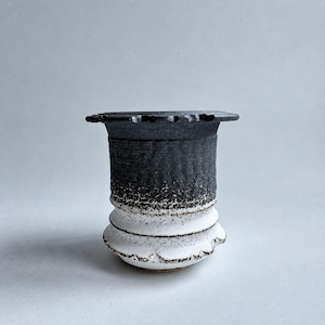 May include: A ceramic vessel featuring a two-tone design with a dark charcoal black upper section and a white lower section. The surface has a rough, textured finish with a wide, flat rim and two distinct horizontal ridges near the base.