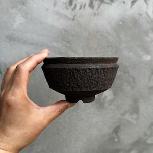 Peut inclure: Un bol en céramique brun foncé et texturé, de forme géométrique unique. Le bol a un bord large et légèrement évasé et une surface texturée et rugueuse. La base a trois petits pieds. Le bol est tenu dans une main sur un fond gris.