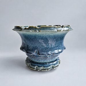May include: A ceramic bowl with a wide, flared rim and a tiered, pedestal-style base. The surface features a mottled glaze in shades of deep navy blue, slate, and hints of light gray, with a glossy finish and a textured, uneven edge along the rim.