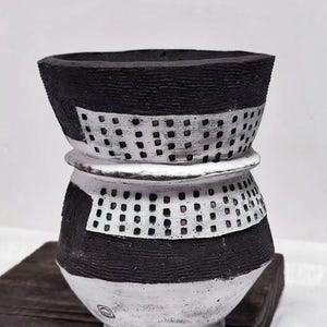 Op de afbeelding: Een handgemaakte keramische vaas met een uniek ontwerp. De vaas heeft een zwart-wit kleurenschema met getextureerde oppervlakken en een geperforeerd patroon. De vaas heeft een brede bovenkant en een afgeronde basis, wat een moderne esthetiek laat zien. Het is een decoratief item.