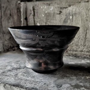 May include: A black ceramic bowl with a wide, flared rim and a tapered base. The surface features a dark, metallic, and textured finish with subtle swirling patterns and a slightly uneven, organic rim edge that creates a distinct silhouette.