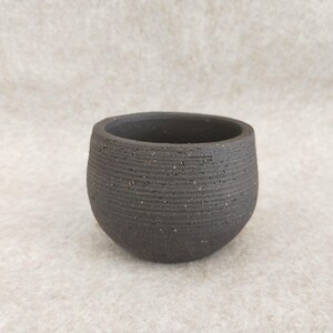 May include: A small, dark gray ceramic bowl with a textured, horizontal-lined pattern. The bowl has a rounded shape and a wide opening. The surface has a speckled appearance. The bowl is likely handmade and suitable for small plants or as a decorative item.