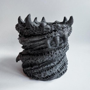 Peut inclure: Un récipient cylindrique gris foncé et texturé sur le thème de la fantaisie. Le récipient a un extérieur rugueux et écailleux et présente des protubérances dentaires et dentelées autour du bord, suggérant la gueule d'une créature. Le design évoque la fantaisie et le macabre.