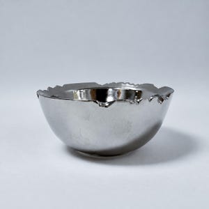 May include: A small, round bowl with a reflective, metallic silver finish. The bowl features a distinct, uneven, and jagged rim that mimics the appearance of a broken or cracked edge, creating a textured look against the smooth, polished surface.