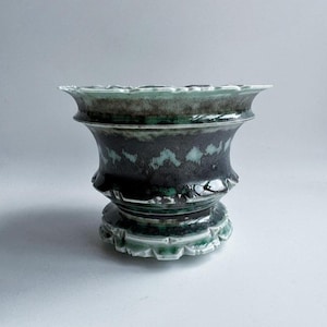 Puede incluir: Un jarrón de cerámica con un borde ancho y festoneado y un cuerpo acampanado. El jarrón presenta un esmalte oscuro y moteado con detalles turquesa y una base decorativa. El diseño recuerda a la cerámica antigua.