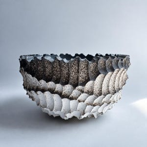 May include: A bowl with a scalloped, wavy rim and a textured, ribbed exterior. The surface features a gradient transition from a dark, charcoal-black interior to a lighter, speckled white base, creating a rhythmic pattern of deep, curved ridges.