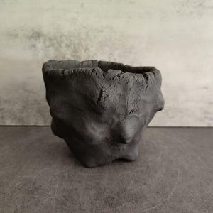 May include: A handmade, dark gray ceramic planter with a rough, textured surface. The small, square-shaped pot has an organic, almost sculptural appearance, perfect for small plants or succulents. The interior is smooth, contrasting with the exterior's rugged look.