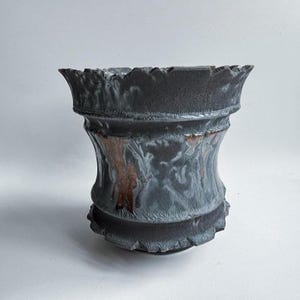 Puede incluir: Un jarrón de cerámica hecho a mano con un exterior gris oscuro texturizado y un borde acampanado y con volantes. El jarrón presenta un diseño único y orgánico con toques de marrón y blanco.
