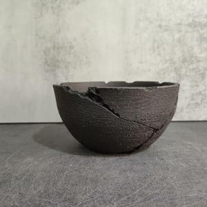 Puede incluir: Un cuenco de cerámica gris oscuro, de forma semiesférica, con una superficie texturizada y una grieta prominente. El borde del cuenco es irregular y el interior es de un tono gris más oscuro. El cuenco se muestra sobre una superficie gris.