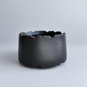 Puede incluir: Un cuenco negro mate de forma cilíndrica ancha con un borde texturizado e irregular que imita la apariencia de bordes de piedra rotos o dentados. La superficie exterior tiene un acabado oscuro y granulado de color negro profundo.