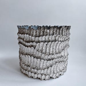 May include: A cylindrical ceramic planter featuring a textured, rippled surface with a high-contrast black and white finish. The exterior displays repeating curved ridges and a speckled, weathered appearance with a dark, scalloped top rim.