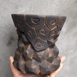 Może przedstawiać: Ciemnobrązowa ceramiczna donica o geometrycznym wzorze. Donica ma unikalny, kanciasty kształt z teksturowaną powierzchnią. Naczynie trzymane jest w dłoni, ukazując jego rozmiar i kształt. Projekt donicy zawiera powtarzające się kształty geometryczne.