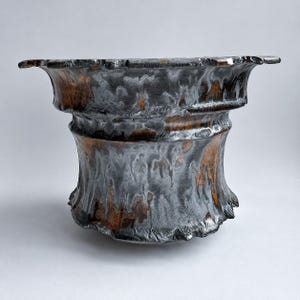 Puede incluir: Un cuenco de cerámica de boca ancha con borde acampanado y base estrecha. La superficie exterior presenta un esmalte moteado con tonos gris carbón y negro profundo que se mezclan con reflejos de color cobre y bronce, creando una textura.