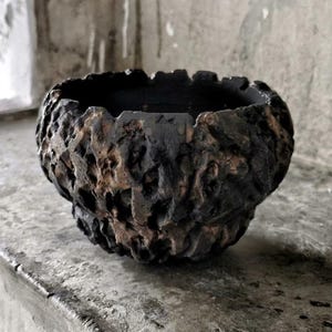 May include: A dark brown and black ceramic bowl with a textured, uneven surface. The bowl has a wide opening and a rounded shape, suitable for decorative use or as a small planter. The interior is a dark, matte black.