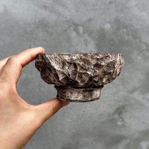 Peut inclure: Un bol en céramique marron texturé avec une base en piédestal. Le bol a une surface rugueuse et inégale et une couleur marron foncé avec des accents blancs. Le bol est tenu dans une main sur un fond gris. Le bol est probablement utilisé pour servir des aliments ou comme objet décoratif.