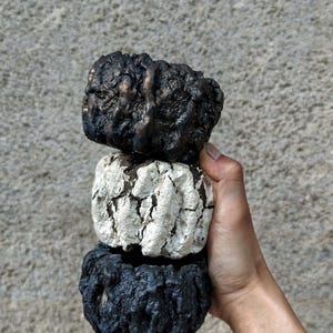 May include: Three small, textured, handmade planters in a stack. The top planter is black with bronze highlights, the middle one is white with dark cracks, and the bottom is black. The planters have a rough, organic appearance.