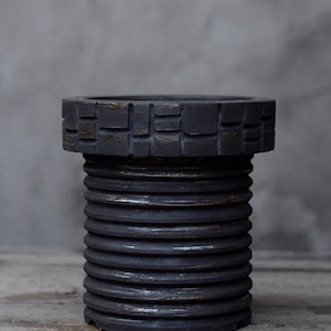 Könnte beinhalten: Ein dunkelgrauer, zylindrischer Behälter mit einem strukturierten, geriffelten Körper und einem dekorativen, quadratisch gemusterten Rand. Der Behälter besteht aus einem dunklen Material und steht auf einer Holzoberfläche. Das Design deutet auf eine rustikale oder industrielle Ästhetik hin.