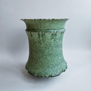 Könnte beinhalten: Eine handgefertigte, grüne Keramikvase mit strukturierter Oberfläche. Die Vase hat eine ausgestellte Ober- und Unterseite mit einem zylindrischen Mittelteil. Der Rand und die Basis haben einen leicht unebenen, organischen Rand. Die Vase ist etwa 20 cm hoch.