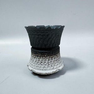 May include: A two-piece ceramic planter with a textured surface. The top section is black with a scalloped rim, and the bottom section transitions from white to black with a speckled pattern. The planter has a unique, artistic design.