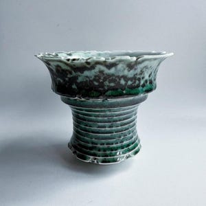 May include: A handcrafted ceramic vase with a distinctive design. The vase has a wide, ruffled rim and a cylindrical base with horizontal grooves. The color scheme features shades of teal and green, creating a visually appealing contrast. The vase is a decorative item.