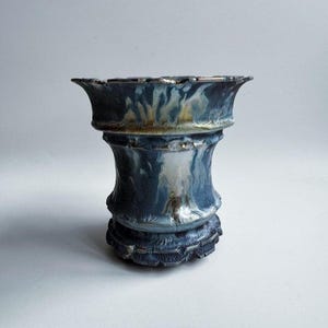 以下が含まれることがあります： ユニークなレイヤードデザインの手作りセラミック花瓶。花瓶は、銀と金のアクセントが付いた濃い青色の釉薬と、テクスチャ加工された溝付きベースが特徴です。縁はスカラップエッジです。花瓶の高さは約20cmです。