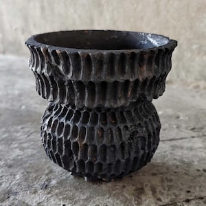 Puede incluir: Un recipiente oscuro y texturizado, hecho a mano, con un diseño orgánico único. La maceta negra presenta una base bulbosa y un borde acampanado, con un patrón tallado de pequeños agujeros irregulares. La superficie tiene una calidad rugosa y táctil.