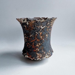 Puede incluir: Un jarrón de cerámica hecho a mano con una superficie texturizada única. El jarrón presenta un borde acampanado y con volantes y una combinación de colores oscuros y moteados con tonos de negro, marrón y blanco. El jarrón tiene una estética rústica y orgánica.