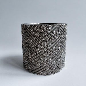 Könnte beinhalten: Ein zylindrisches Gefäß aus silberfarbenem Metall mit einem tief geprägten geometrischen Muster aus sich kreuzenden Linien und kantigen Formen. Die Oberfläche hat ein reflektierendes, poliertes Finish mit hohem Kontrast.
