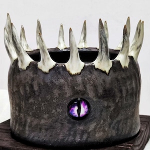 Puede incluir: Un cuenco de cerámica gris oscuro con una corona de púas afiladas con puntas blancas. El cuenco presenta un adorno en forma de ojo morado y negro. El cuenco está sobre una base de madera oscura, creando una pieza decorativa única.
