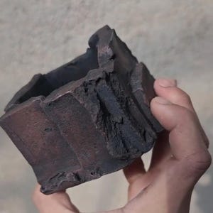 May include: A dark brown, square-shaped ceramic planter with a rough, textured surface. The planter has a unique, organic design with uneven edges and a dark, earthy color. It is held in a hand.