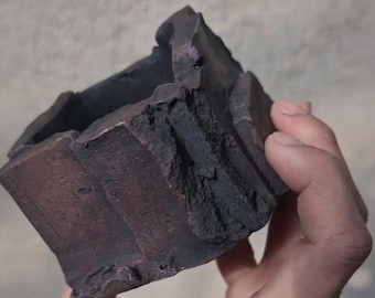 Op de afbeelding: Een donkerbruine, vierkante keramische plantenbak met een ruw, getextureerd oppervlak. De plantenbak heeft een uniek, organisch ontwerp met ongelijke randen en een donkere, aardse kleur. Hij wordt in een hand gehouden.