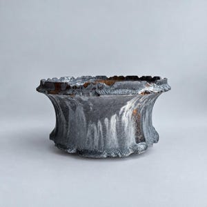 May include: A wide, low-profile ceramic bowl featuring a textured, dark grey and charcoal exterior with vertical, streaked patterns. The rim is uneven and jagged, revealing hints of warm brown and amber tones along the inner edge and top surface.