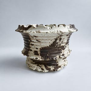 May include: A ceramic planter featuring a flared, jagged rim and a ribbed body. The surface is finished with a speckled pattern of dark brown and off-white glaze, creating a high-contrast appearance across the entire exterior of the vessel.