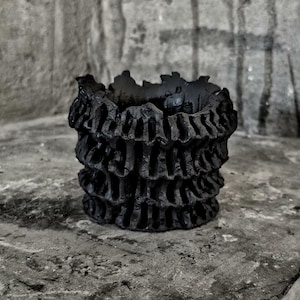 May include: A black, textured pot with a distinctive layered design. The pot has a rough, organic appearance, with stacked, openwork tiers. The dark color and intricate details give it a modern, artistic feel. Suitable for home decor.